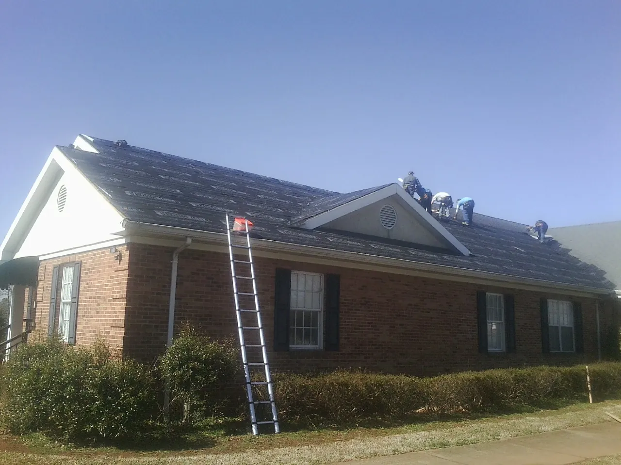 Crew tearing shingles off a brick ranch roof with a ladder set — Emergency Roof Repair work by our crew in Riley