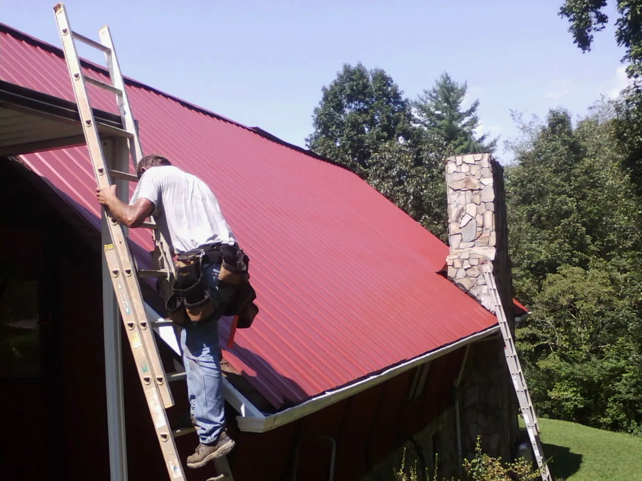 Roofer on a ladder beside a red metal roof and stone chimney — Soffit & Fascia Repair by Roofing Brothers in Riley
