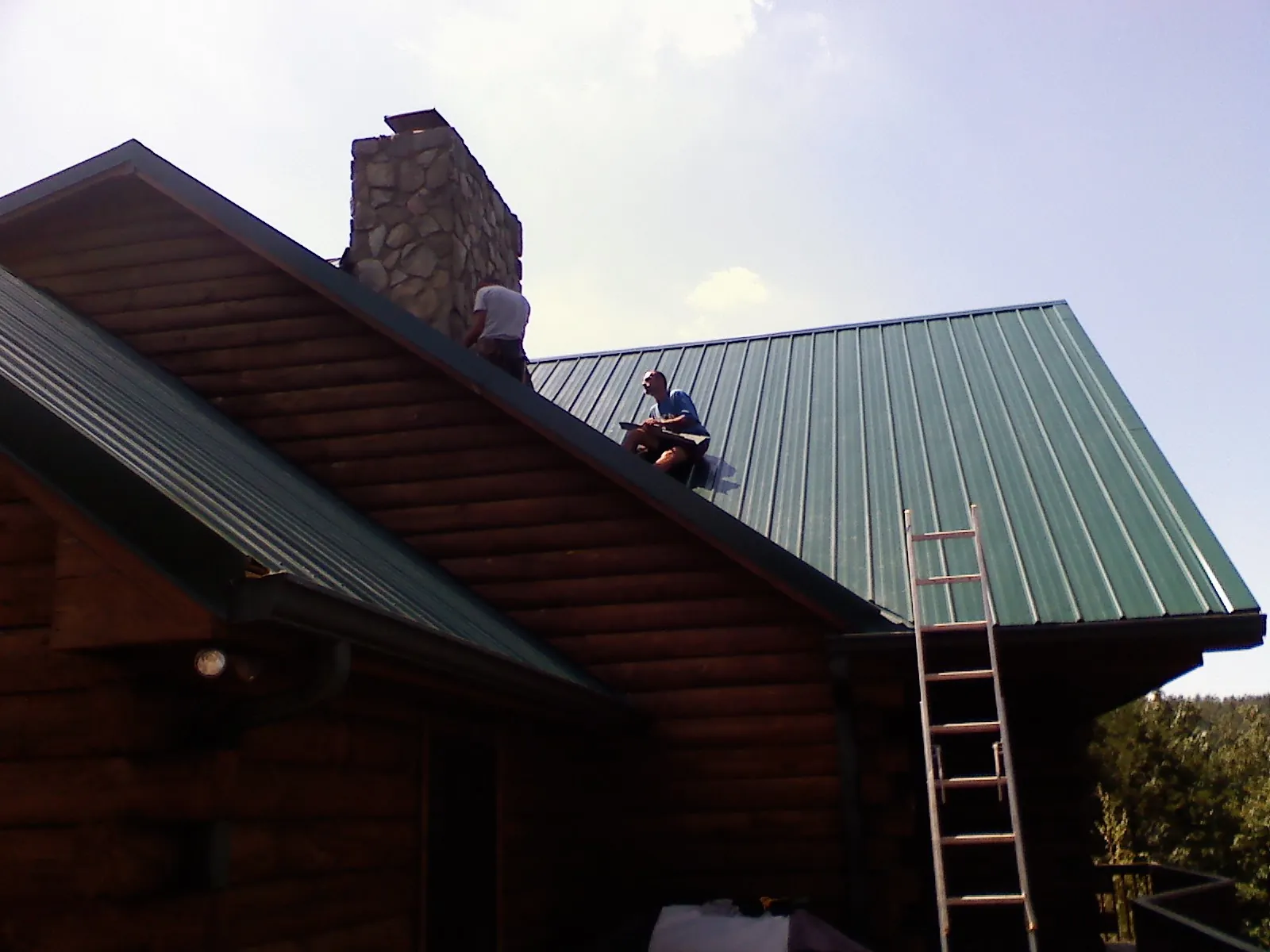 Roofers installing green metal panels beside a stone chimney on a log home in Riley